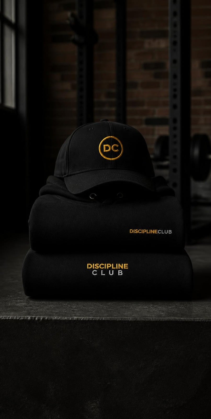 Discipline Club Hoodie and Crewneck folded and stacked with the Crest Structured Cap on a concrete bench in the gym